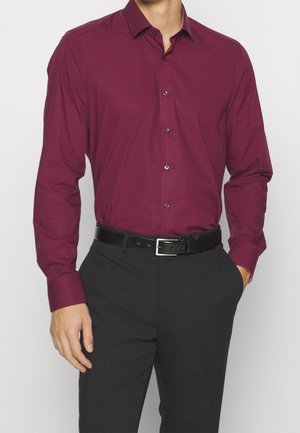 Homme portant une chemise bordeaux à manches longues rentrée dans un pantalon noir avec une ceinture noire, debout avec une main dans la poche.
