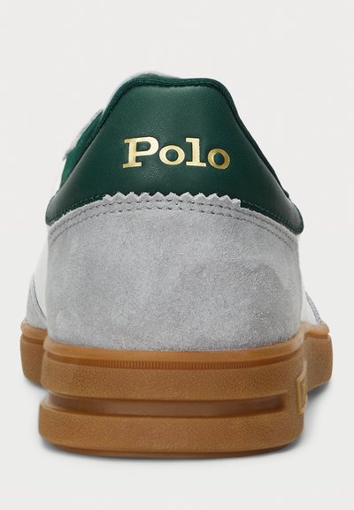 Sivi nubuk športni copat z zelenim usnjenim petnim delom, zlatim logotipom "Polo" in gumijastim podplatom. Ima okroglo obliko in teksturirano površino.