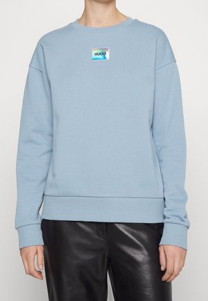 Person trägt einen hellblauen Pullover mit Rundhalsausschnitt und einem kleinen holografischen "HUGO"-Logo auf der Brust, kombiniert mit schwarzen Lederhosen.