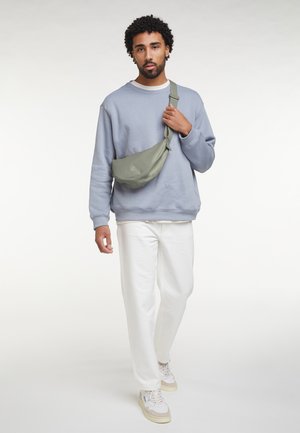 Man draagt een lichtblauwe trui, witte broek en sneakers, en draagt een olijfgroene schoudertas terwijl hij vooruit loopt.