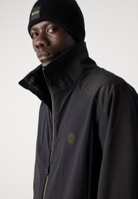 Black zip-up jacket with high collar, featuring a textured logo patch. Accompanied by a black knitted beanie with a logo tag.