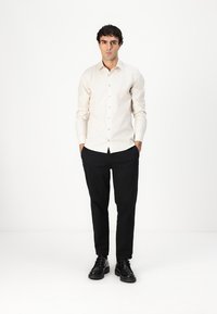 Lys beige knappet skjorte med en skarp krage, kombinert med tettsittende svarte bukser og svarte sko, på en modell som står mot en hvit bakgrunn.