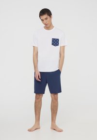T-shirt bianco con una tasca blu decorata con motivi di palme, abbinato a pantaloni corti navy. L'outfit è semplice e informale.