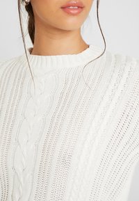 Young woman wearing large silver hoop earrings and a white cable-knit sweater with vertical patterns.