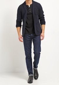 Blazer a maglia blu navy con tasca, indossato sopra una maglietta nera a manica lunga. Abbinato a jeans blu scuro e stivaletti neri alla caviglia. Look casual.