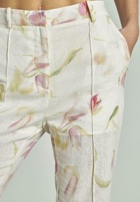 Close-up van witte broeken met een zacht roze en groene bloemenprint, featuring zijzakken en een tailleband met riemlussen.