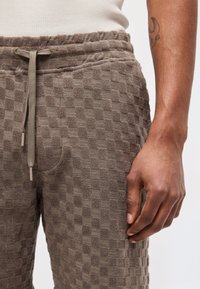 Gros plan sur une personne portant un pantalon marron à carreaux texturé avec une taille élastique et un cordon de serrage, la main reposant à côté.