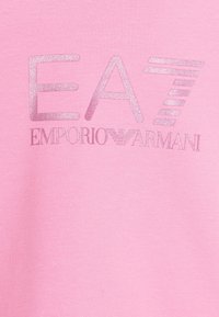 Różowy materiał z błyszczącym srebrnym napisem "EA7" oraz logo "EMPORIO ARMANI" w stylizowanej czcionce, z emblematem poniżej liter.