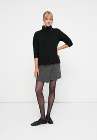 Czarny sweter z golfem, szara spódnica w kratę, przezroczyste czarne rajstopy i czarne płaskie buty w minimalistycznym otoczeniu.