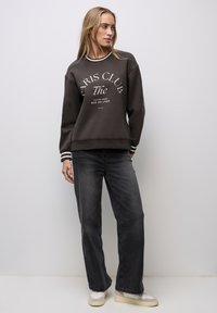 Sweatshirt gris foncé avec impression "Paris Club", texte blanc, poignets rayés, associé à un jean noir à jambes larges et des sneakers claires.