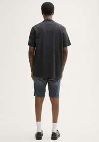 Cămașă neagră cu mâneci scurte și design texturat, asociată cu pantaloni scurți din denim albastru, șosete albe și pantofi negri, văzută din spate.