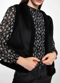 Svart ärmlös väst med ett mjukt, texturerat kantband. Under är en svart blus med blommönster, lång puffade ärmar och hög halsringning.