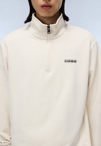 Langärmliger Pullover in Cremefarbe mit halbem Reißverschluss am Kragen. Verfügt über einen Logo-Patch auf der Brust und eine weiche, glatte Stoffstruktur.