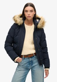 Doudoune marine avec capuche doublée en fausse fourrure, zip sur le devant et deux poches latérales, associée à un pull crème en maille torsadée et un jean bleu.