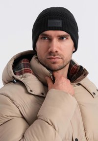 Black knitted beanie with a textured pattern and a rectangular logo patch. Beige puffer jacket with plaid lining, shown partially zipped.