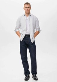 Camisa de botones a rayas blancas y azules sobre una camiseta blanca, combinada con vaqueros azul oscuro y zapatos negros, de pie contra un fondo liso.