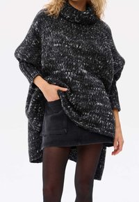 Schwarz-weiß gepunkteter Strickponcho mit hohem Kragen und lässiger Passform. Kombiniert mit einem dunkelblauen Rock aus Denim und schwarzen Strumpfhosen.