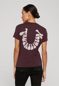 Žena s tmavými vlasy v drdolu, která má na sobě fialové tričko se stylizovaným bílým glitrovým textem tvořícím tvar úsměvu na zádech, doplněné černými kalhotami.