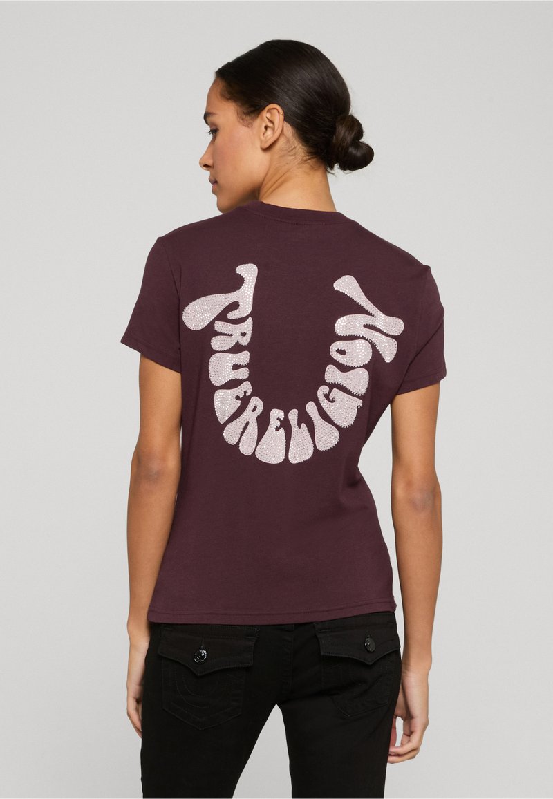 Žena s tmavými vlasy v drdolu, která má na sobě fialové tričko se stylizovaným bílým glitrovým textem tvořícím tvar úsměvu na zádech, doplněné černými kalhotami.