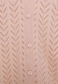 Ljusrosa stickad cardigans med ett zigzag-mönster och knappstängning. Texturen är fin och luftig, vilket visar på detaljerat hantverk.