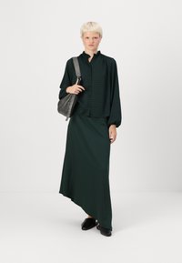 Tamsiai žalia bluzonė su tekstūriniais raštais, derinama su asimetriška tamsiai žalia sijonu. Modelis laiko pilką rankinę, vilki juodus batus.