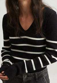 Schwarz-weißer gestreifter gerippter Pullover mit V-Ausschnitt, langärmlig und mit eng anliegenden Bündchen. Getragen mit dunklen Jeans.