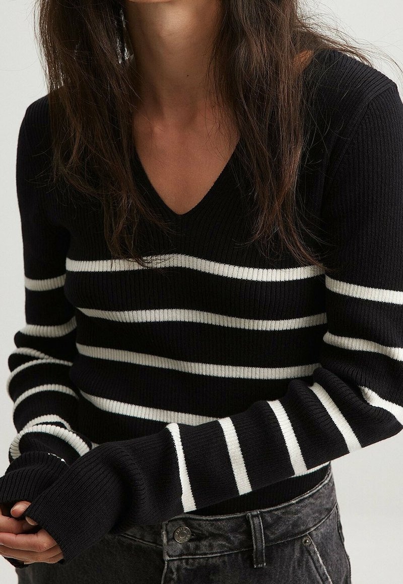 Schwarz-weißer gestreifter gerippter Pullover mit V-Ausschnitt, langärmlig und mit eng anliegenden Bündchen. Getragen mit dunklen Jeans.
