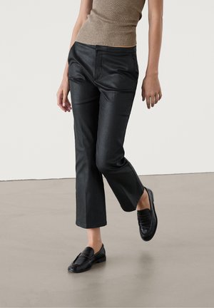 Osoba ubrana w krótkie czarne skórzane spodnie, beżowy bezrękawnik i czarne mokasyny, stojąca na gładkiej szarej podłodze.