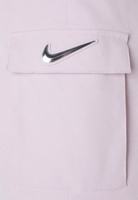 Svetle fialová látka s hladkým povrchem, sešitá kapsa a kovové logo Nike na klopě. Jednoduchý design a funkční detail.