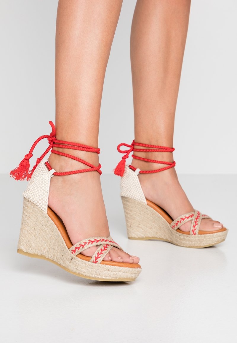 Pieds portant des sandales compensées beige avec des brides tressées rouges nouées autour des chevilles et attachées autour des jambes inférieures sur un fond clair.