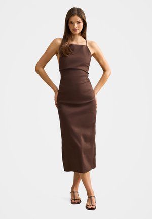 Žena stojaca s rukami v bok, oblečená v priliehavých hnedých midi šatách bez rukávov a čiernych sandáloch na podpätku s remienkami.