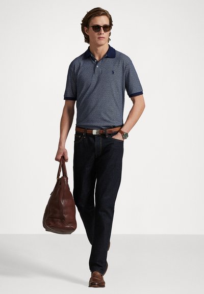 Polo azul-marinho com design padr�ão, jeans escuros, cinto de couro castanho e óculos de sol. A transportar uma mala de couro castanho. Corte clássico.