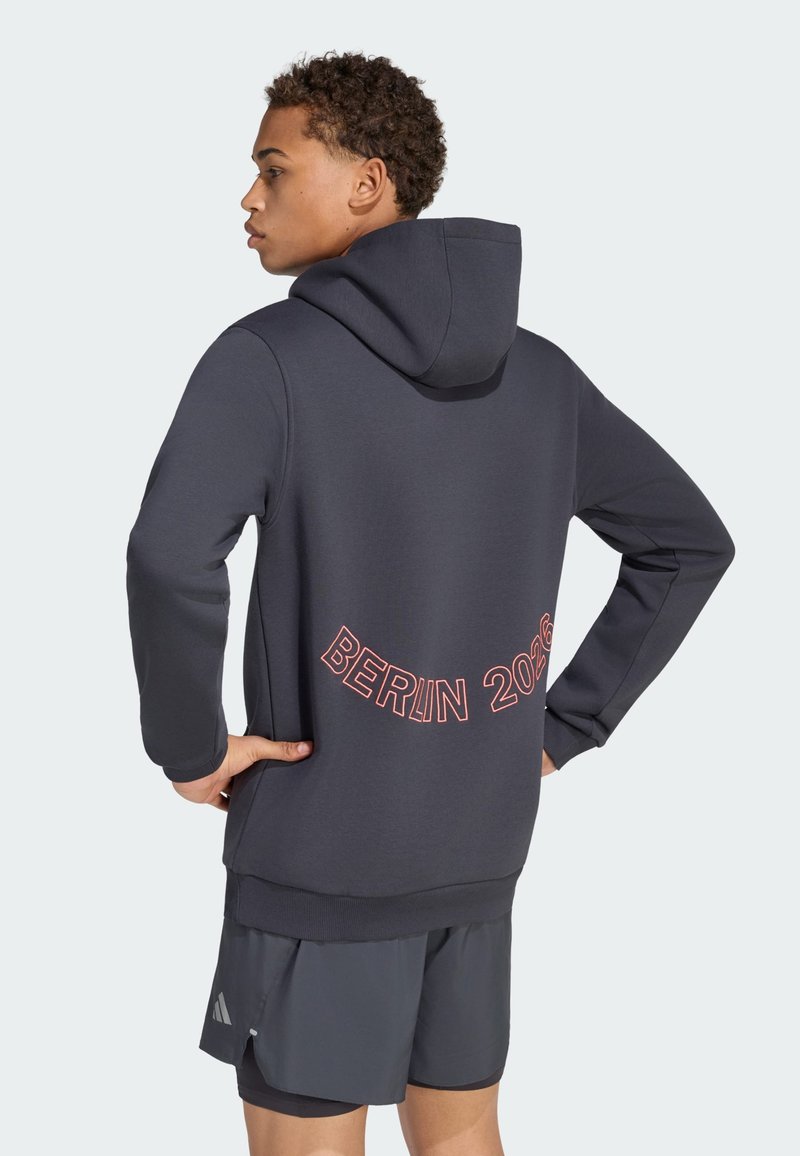 Jeune homme portant un sweat à capuche foncé avec « BERLIN 2026 » imprimé dans le dos, assorti à un short de sport foncé.