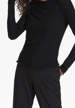 Persoon draagt een nauwsluitend zwart shirt met lange mouwen en gerimpelde zoom en losse zwarte broek, poseert met één hand op het hoofd en de andere in de zak.