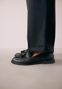 Sorte læder loafers med frynseaccent, glat tekstur, rund tå og lav hæl, parret med skræddersyede sorte bukser.