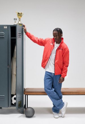 Rød vindjakke, hvid T-shirt, blå jeans og hvide sneakers. Skab med trofæer, fodbold og grå basketball i nærheden.