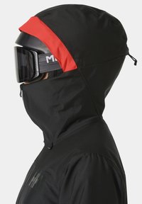 Černá lyžařská bunda s červeným akcentem na kapuci, s vysokým límcem, zipy a texturovaným povrchem. Obsahuje helmu s kšiltem a brýle.