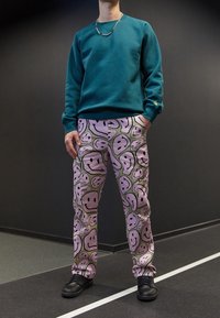 Sudadera azul verdosa con cuello redondo y puños acanalados, combinada con pantalones rosas cubiertos de ilustraciones de caritas sonrientes negras. Zapatos negros de diseño minimalista.