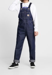 Mörkblå denimöveraller med justerbara axelremmar, framficka, guldfärgad hårdvara och upprullade ärmslut. Åt en svart sneakers.