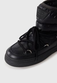Black winter boot with a puffy upper, lace closure, silver hardware accents, textured black rubber sole, and a branded patch near the top.