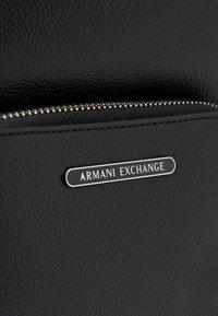 Juodas tekstūruotas sintetinės medžiagos paviršius su užtrauktuku. Pasižymi metaliniu "Armani Exchange" logotipu. Lygus paviršius su smulkmenomis.