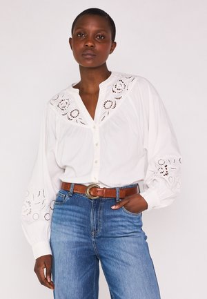 Donna che indossa una camicetta bianca con ricamo floreale traforato sul colletto e sulle maniche, jeans blu e una cintura marrone con fibbia dorata.