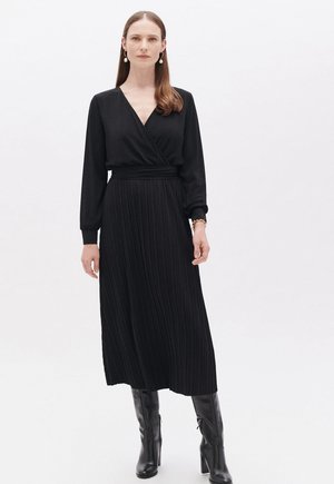Abito nero a maniche lunghe con stile avvolgente, gonna plissettata e vita elasticizzata. Indossato con stivali neri al ginocchio.