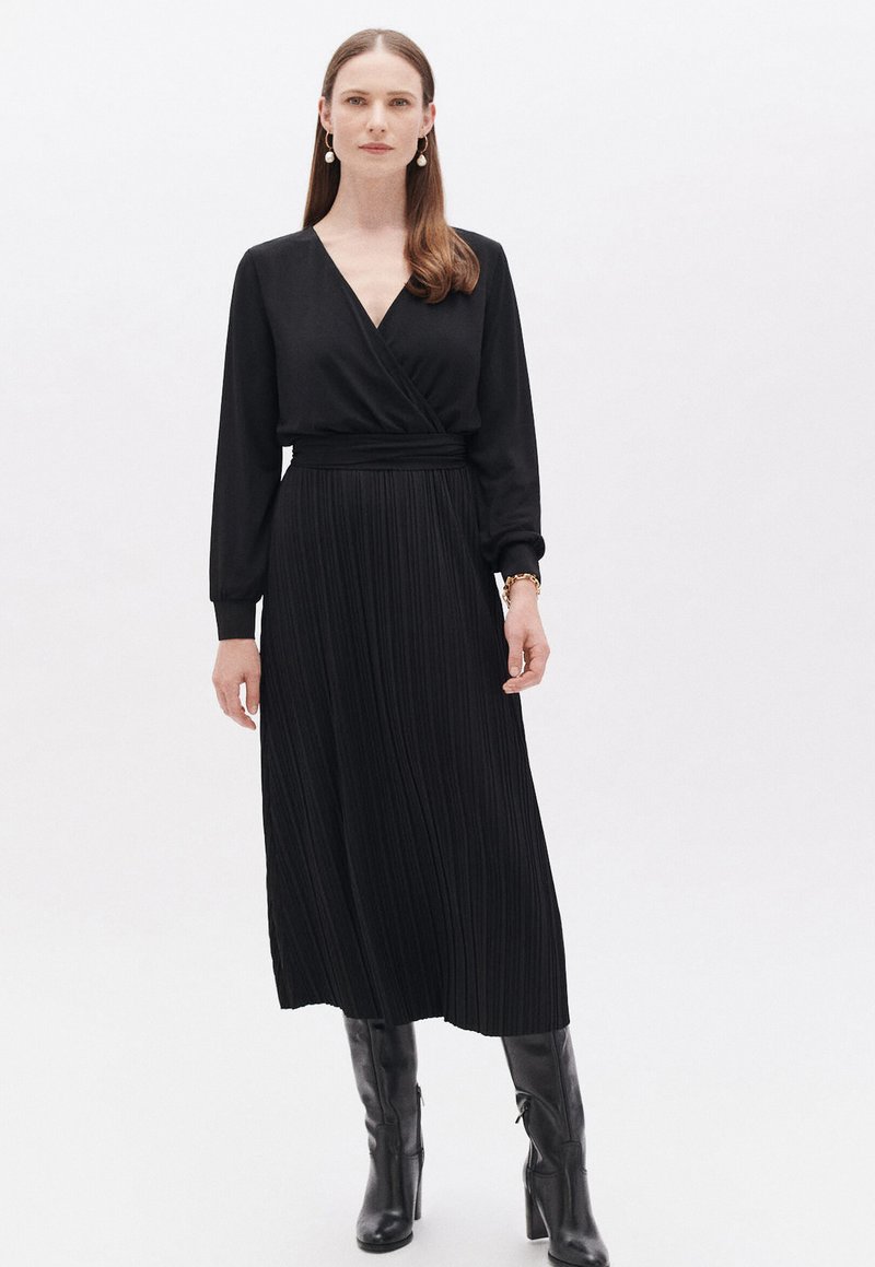 Schwarzes langärmeliges Kleid im Wickelstil, mit plissiertem Rock und elastischem Bund. Getragen mit knielangen schwarzen Stiefeln.