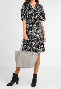 Sac tote en cuir gris avec double poignées, présentant une texture lisse et une forme rectangulaire, à côté d'une robe à motifs noir et blanc.