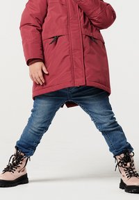 Abrigo aislante rojo con dos bolsillos frontales y detalles en negro en la cremallera, combinado con jeans de mezclilla azules y botas rosas con cordones y suelas negras.