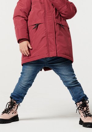 Červený zateplený kabát s dvěma předními kapsami a černými zipovými akcenty, spárovaný s modrými denimovými džíny a růžovými šněrovacími botami s černými podrážkami.