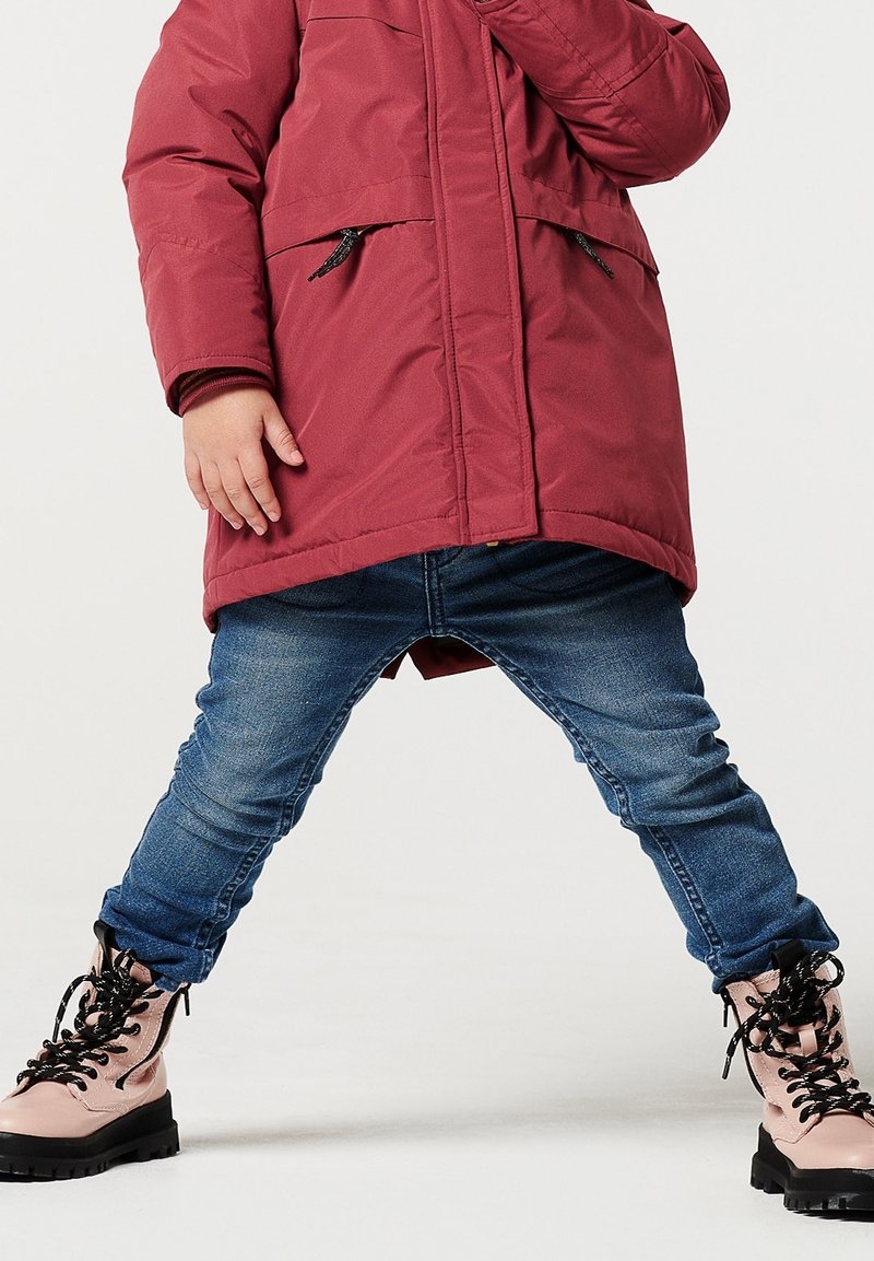 Abrigo aislante rojo con dos bolsillos frontales y detalles en negro en la cremallera, combinado con jeans de mezclilla azules y botas rosas con cordones y suelas negras.