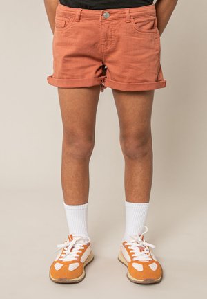 Personne portant un short orange retroussé, des chaussettes blanches mi-mollet et des baskets orange et blanches, debout devant un fond neutre.