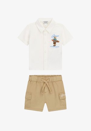 Chemise blanche à manches courtes avec boutons, ornée d'un motif d'ours en peluche et de planche de surf, associée à un short beige à cordon de serrage avec poches latérales et logo Guess.